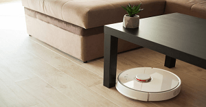 Robot vacuum cleaner cleaning a wooden floor near a black coffee table and a beige sofa.