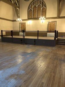 A large, empty hall with a raised stage. Wooden floor and walls, ornate arched window boarded up.