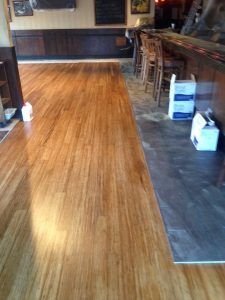 Hardwood floor in a bar extends to concrete floor near bar counter, chairs, and boxes.